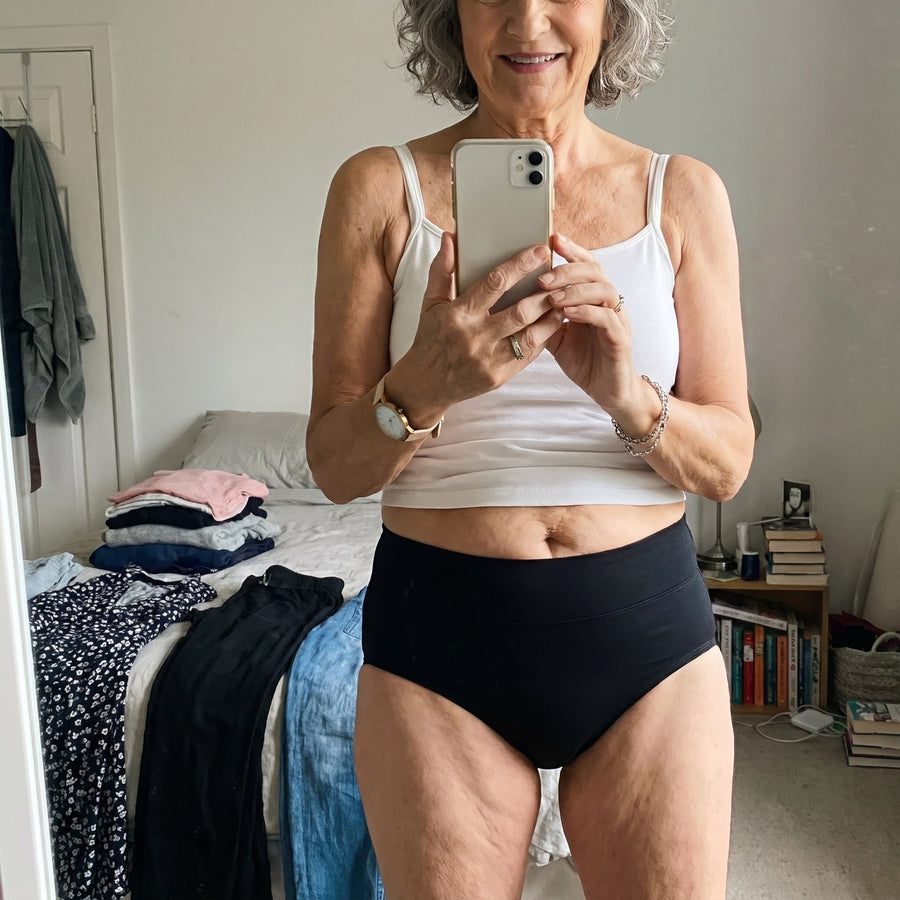 Donna sulla sessantina, sorridente, che si scatta un selfie allo specchio con canotta bianca e lo slip lavabile Fresia nero a vita alta